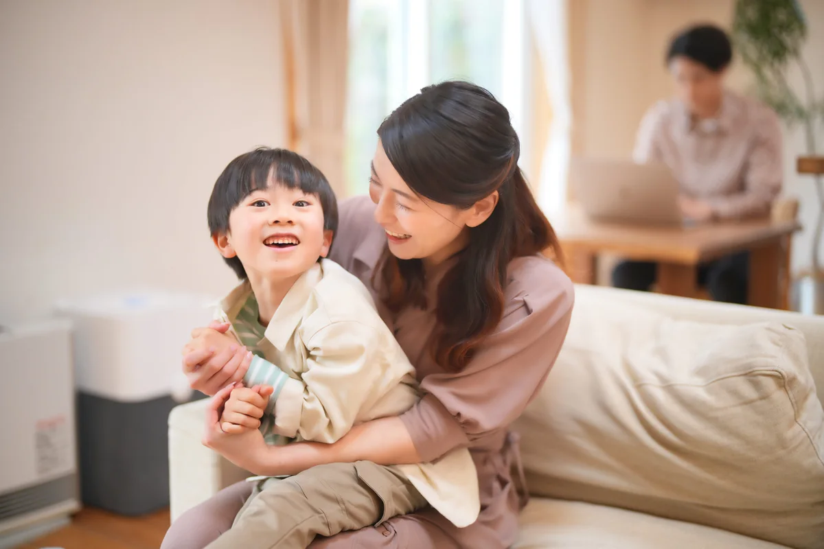 リビングで子どもを抱く母親と、奥でパソコンを使う父親